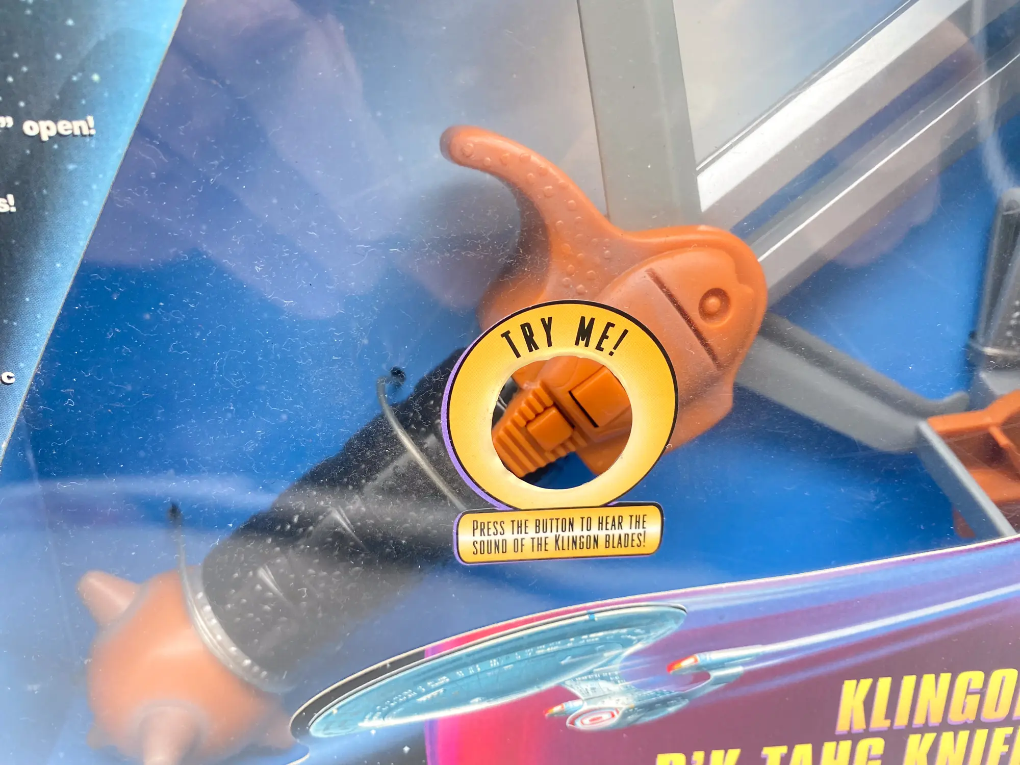 Gros plan sur le bouton sonore et la garde orange du Klingon D’k Tahg, derrière la fenêtre marquée.